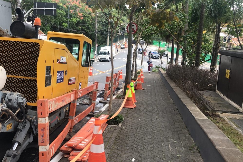 Perfuratriz da DRC operando na calçada atrás de barreiras, com a calçada liberada para pedestres e a avenida com tráfego normal ao fundo
