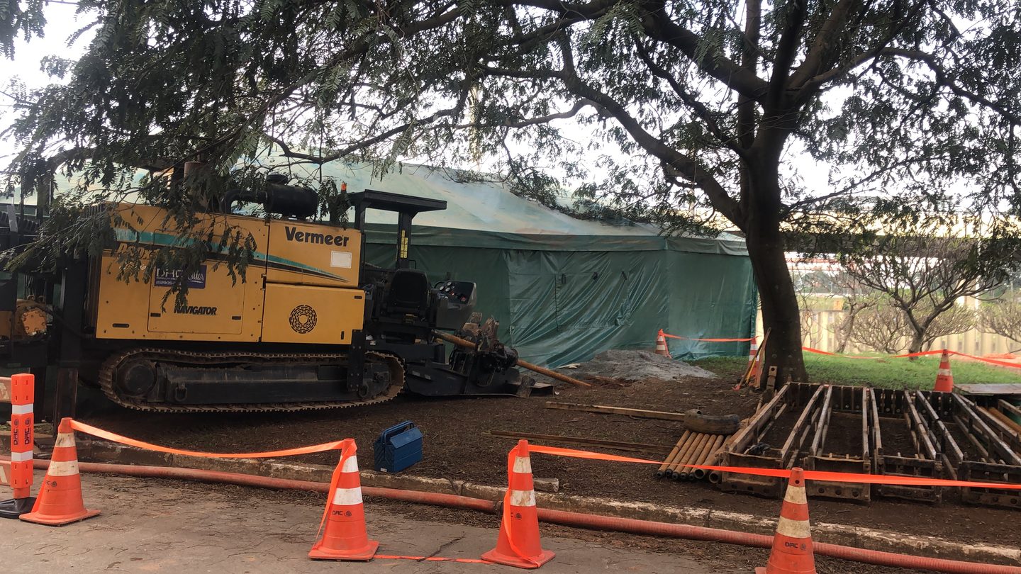 Perfuratriz direcional Vermeer da DRC em frente de obra compacta, isolada por cones da própria empresa, com hastes de perfuração ao lado e árvore preservada sobre o canteiro